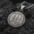 Load image into Gallery viewer, Oxidized Tiberius coin pendant necklace resting on black volcanic stone, featuring a Roman emperor profile and antique coin-style inscription in a detailed macro product photograph.