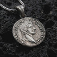 Load image into Gallery viewer, Oxidized Tiberius coin pendant necklace resting on black volcanic stone, featuring a Roman emperor profile and antique coin-style inscription in a detailed macro product photograph.
