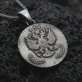 Load image into Gallery viewer, Oxidized heraldic eagle coin pendant necklace resting on black volcanic stone, featuring a round medallion with a crowned double-headed eagle, stars, and laurel branches in a detailed macro product photograph.