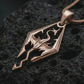 Load image into Gallery viewer, Rose gold plated Skyrim dragon pendant necklace on textured volcanic rock, detailed macro product shot with soft warm reflections and bold dragon emblem styling.