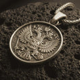 Load image into Gallery viewer, Bronze double headed eagle pendant necklace resting on black volcanic stone, featuring a detailed heraldic eagle crest with spread wings and central shield in a macro product photograph.