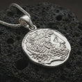 Load image into Gallery viewer, Polished silver ancient Greek coin pendant necklace displayed on dark volcanic rock, shown in a premium close-up image that highlights the stylized male portrait and organic antique coin texture.