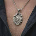Load image into Gallery viewer, Oxidized Saint Padre Pio spinning pendant necklace worn on the neck, styled in a dark lifestyle image that presents the Catholic medal as a meaningful symbol of faith, prayer, and protection.