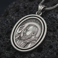 Load image into Gallery viewer, Oxidized Saint Padre Pio spinning pendant necklace resting on black volcanic stone, featuring an oval double-frame medal with a detailed portrait of Padre Pio raising his hand in blessing.