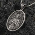 Load image into Gallery viewer, Oxidized Saint Francis pendant necklace on textured volcanic rock, close-up studio shot highlighting the saint relief, bird, wolf detail, and antique silver contrast.
