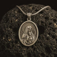 Load image into Gallery viewer, Bronze Virgin Mary and baby Jesus pendant necklace on textured volcanic rock, close-up studio shot highlighting the mother and child relief, halo, star border, and antique silver contrast.