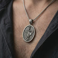 Load image into Gallery viewer, Oxidized Saint Michael pendant necklace worn on the chest, close-up lifestyle jewelry photo showing the archangel medallion on a spiga chain with dark clothing.