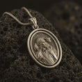 Load image into Gallery viewer, Bronze spinning Good Shepherd pendant necklace on textured volcanic rock, close-up studio shot highlighting the Jesus and lamb relief with antique silver contrast.