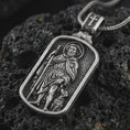Load image into Gallery viewer, Oxidized Saint Roch dog tag pendant necklace on textured volcanic rock, close-up studio shot highlighting the pilgrim saint relief, staff, dog detail, and antique silver contrast.