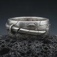 Load image into Gallery viewer, Oxidized Bushido sword band ring on textured volcanic rock, close-up studio shot highlighting the wrapped sword motif, cracked metal texture, rising sun detail, and antique silver contrast.