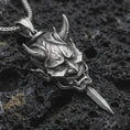 Load image into Gallery viewer, An oxidized oni mask pendant rests on dark volcanic rock in a macro studio close-up, showing a fierce horned demon face with a blade-like detail.