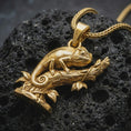 Load image into Gallery viewer, Gold plated chameleon pendant necklace on textured volcanic rock, premium close-up studio composition emphasizing the climbing lizard, curled tail, and nature-inspired branch design.