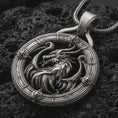 Load image into Gallery viewer, An oxidized dragon pendant rests on dark volcanic rock in a macro studio close-up, showing a fierce mythical creature inside a detailed round frame.
