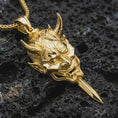 Load image into Gallery viewer, A gold plated oni mask pendant lies on textured volcanic rock in a dramatic studio macro shot, highlighting the protective demon mask design.