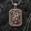 Load image into Gallery viewer, A Bronze Hannya mask pendant rests on dark volcanic rock in a macro studio close-up, showing a fierce Japanese demon face inside an ornate frame.