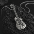 Load image into Gallery viewer, Oxidized skull guitar pendant necklace on textured volcanic rock, close-up studio shot highlighting the guitar body, skull detail, rock hand gesture, music notes, and antique silver contrast.