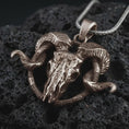 Load image into Gallery viewer, Bronze ram skull pendant necklace on textured volcanic rock, close-up studio shot highlighting the cracked skull, curled horns, skeletal details, and antique silver contrast.