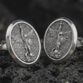Load image into Gallery viewer, Oxidized Saint Michael cufflinks on textured volcanic rock, close-up studio shot highlighting the archangel relief, spear, wings, patterned background, and antique silver contrast.