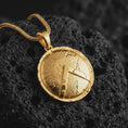 Load image into Gallery viewer, Gold plated Spartan shield pendant necklace on textured volcanic rock, premium close-up studio composition emphasizing the ancient Greek shield, lambda emblem, and rugged cracked surface.