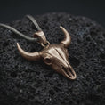 Load image into Gallery viewer, Bronze bull skull pendant necklace on textured volcanic rock, close-up studio shot highlighting the long horns, hollow skull form, western design, and antique silver contrast.