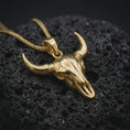 Load image into Gallery viewer, Gold plated bull skull pendant necklace on textured volcanic rock, premium close-up studio composition emphasizing the western skull silhouette, curved horns, and rugged amulet design.