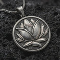 Load image into Gallery viewer, Oxidized lotus flower pendant necklace on textured volcanic rock, close-up studio shot highlighting the layered petals, round medallion frame, textured background, and antique silver contrast.