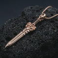 Load image into Gallery viewer, A rose gold plated fantasy sword pendant is photographed on porous volcanic rock in a close-up studio composition with bold luxury lighting and detailed blade texture.