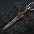Load image into Gallery viewer, A Bronze fantasy sword pendant rests on dark volcanic rock in a macro studio close-up, showing a detailed warrior blade with gothic sculptural elements.