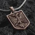 Load image into Gallery viewer, Bronze double-headed eagle shield pendant necklace on textured volcanic rock, close-up studio shot highlighting the twin eagle heads, spread wings, crest frame, and antique silver contrast.