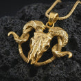 Load image into Gallery viewer, Gold plated ram skull pendant necklace on textured volcanic rock, premium close-up studio composition emphasizing the gothic skull shape, spiral horns, and dark Viking-inspired symbolism.