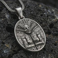 Load image into Gallery viewer, An oxidized Aztec mask pendant rests on dark volcanic rock in a macro studio close-up, showing an ancient warrior face with detailed tribal patterns.
