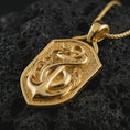 Load image into Gallery viewer, Gold plated serpent and anchor pendant necklace on textured volcanic rock, premium close-up studio composition emphasizing the protective snake, anchor symbol, and nautical shield design.