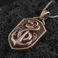 Load image into Gallery viewer, Bronze serpent and anchor pendant necklace on textured volcanic rock, close-up studio shot highlighting the coiled snake, nautical anchor, shield frame, and antique silver contrast.