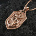 Load image into Gallery viewer, Rose gold plated serpent and anchor pendant necklace on textured volcanic rock, detailed macro product shot with soft warm reflections and maritime amulet symbolism.