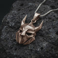 Load image into Gallery viewer, An Bronze Loki pendant rests on dark volcanic rock in a macro studio close-up, showing a faceted horned mask inspired by Norse mythology.