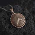 Load image into Gallery viewer, Bronze Spartan shield pendant necklace on textured volcanic rock, close-up studio shot highlighting the cracked round shield, raised lambda symbol, battle-worn texture, and antique silver contrast.