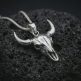Load image into Gallery viewer, Polished silver bull skull pendant necklace on textured volcanic rock, centered macro jewelry photo with detailed horn structure, skull texture, and bright silver highlights.