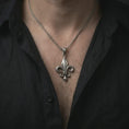Load image into Gallery viewer, Oxidized fleur-de-lis pendant necklace worn on the chest, close-up lifestyle jewelry photo showing the medieval heraldic charm on a spiga chain with dark clothing.