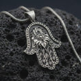 Load image into Gallery viewer, Oxidized hamsa hand pendant necklace on textured volcanic rock, close-up studio shot highlighting the evil eye center, lotus flower relief, ornate hand shape, and antique silver contrast.