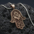 Load image into Gallery viewer, Bronze hamsa hand pendant necklace on textured volcanic rock, close-up studio shot highlighting the evil eye center, lotus flower relief, ornate hand shape, and antique silver contrast.