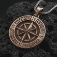Load image into Gallery viewer, Bronze Greek compass pendant necklace on textured volcanic rock, close-up studio shot highlighting the raised compass rose, north south letters, Greek key border, and antique silver contrast.