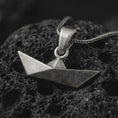 Load image into Gallery viewer, Oxidized origami boat pendant necklace on textured volcanic rock, close-up studio shot highlighting the folded paper boat shape, geometric angles, brushed surface, and antique silver contrast.