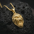 Load image into Gallery viewer, A gold plated gorilla pendant lies on textured volcanic rock in a dramatic studio macro shot, highlighting the powerful ape head design and primal expression.