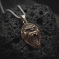 Load image into Gallery viewer, A bronze gorilla pendant rests on dark volcanic rock in a macro studio close-up, showing a fierce roaring ape head with detailed teeth and fur texture.