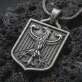 Load image into Gallery viewer, Oxidized German eagle shield pendant necklace on textured volcanic rock, close-up studio shot highlighting the heraldic eagle relief, shield frame, feather details, and antique silver contrast.