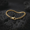 Load image into Gallery viewer, Gold plated men’s box chain bracelet resting across dark volcanic rock, with its narrow geometric link pattern and polished lobster clasp captured in a luxury macro studio close-up.