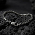 Load image into Gallery viewer, Slim sterling silver box chain bracelet with an oxidized finish arranged in a curved shape on black volcanic rock, showing its compact geometric links and lobster clasp in a macro studio close-up.
