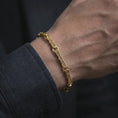 Load image into Gallery viewer, Gold plated Greek key bracelet worn on a man’s wrist with dark formal clothing, highlighting the continuous symbolic pattern in a premium masculine lifestyle photograph.