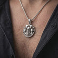 Load image into Gallery viewer, The polished silver Japanese samurai pendant is worn on a man’s chest, with the detailed helmet, crossed swords, armor plates, and sculpted warrior face clearly visible in a premium lifestyle photograph.