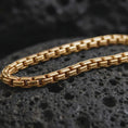 Load image into Gallery viewer, Gold plated box chain arranged across black volcanic rock, emphasizing its squared link construction and bold linear texture.